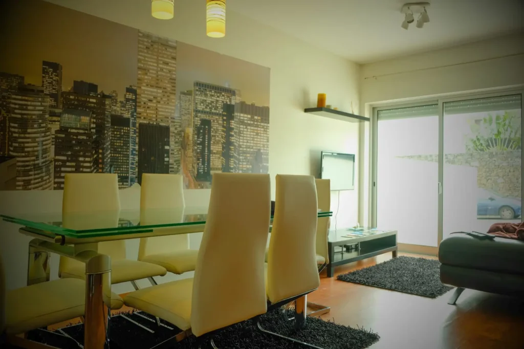 EN: Open-plan living area at Guest House da Fajã de Baixo featuring a glass dining table with high-back white chairs, a modern city skyline mural, and a lounge area with a sofa and TV. PT: Área de estar em plano aberto na Guest House da Fajã de Baixo com mesa de jantar em vidro, cadeiras brancas de encosto alto, mural de parede com skyline de cidade e zona de estar com sofá e TV.