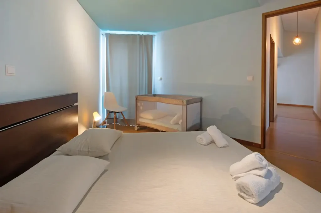 EN: Family bedroom at Ponta Delgada Apartment T3 featuring a double bed with white linens, a complimentary baby crib, a white chair, and a view into the hallway. PT: Quarto familiar no Ponta Delgada Apartment T3 com cama de casal, roupa de cama branca, berço de bebé gratuito, cadeira branca e vista para o corredor.