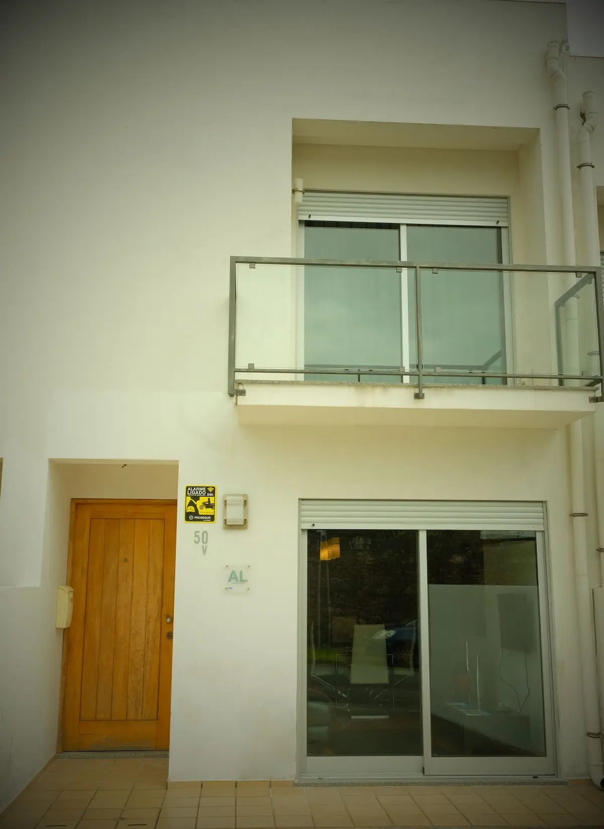 Modern white facade of Guest House da Fajã de Baixo featuring a wooden entrance door, glass balcony, and large sliding windows in Ponta Delgada. PT: Fachada moderna e branca da Guest House da Fajã de Baixo com porta de entrada em madeira, varanda de vidro e grandes janelas de correr em Ponta Delgada.