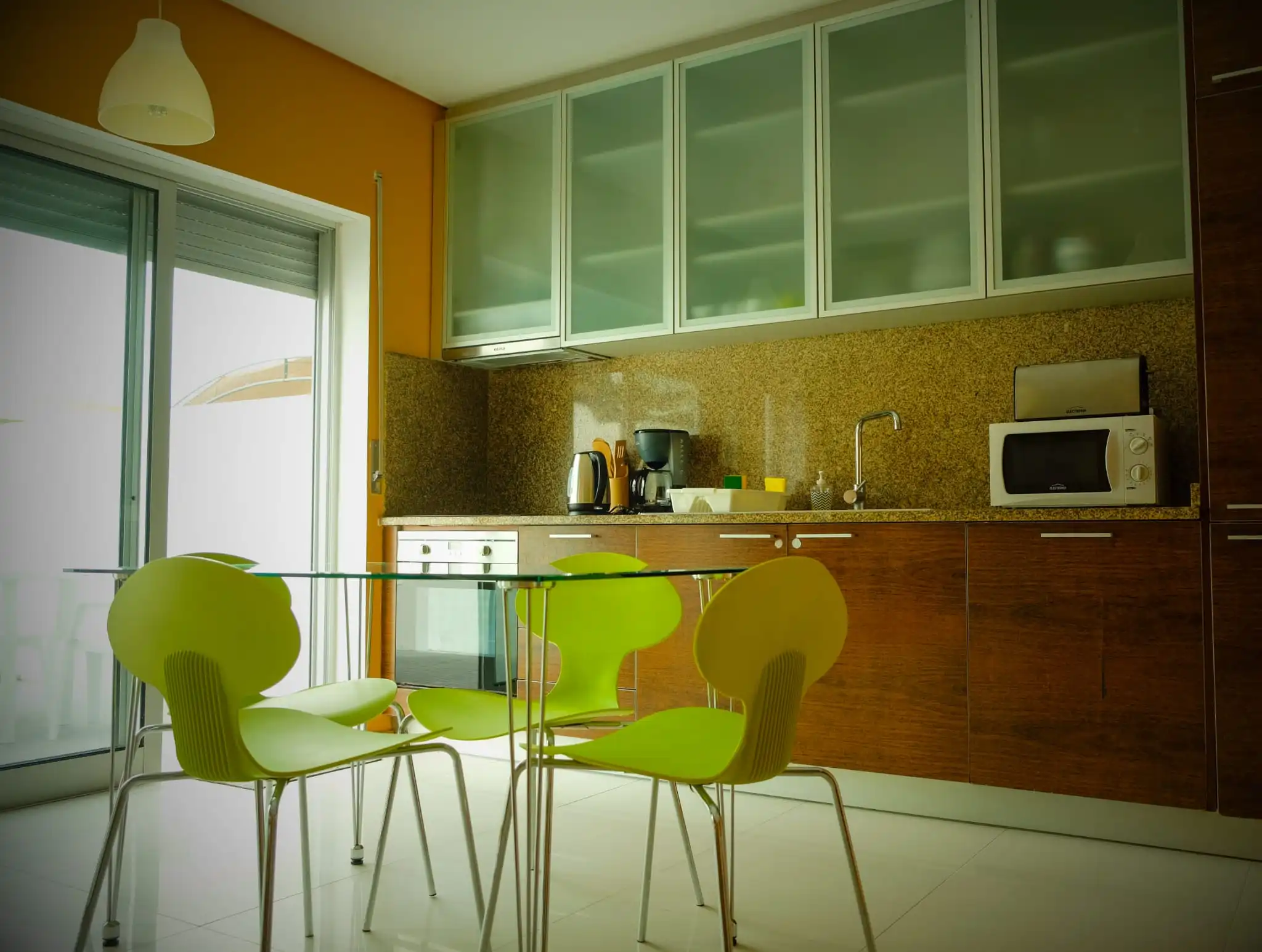 Fully equipped modern kitchen with wooden cabinets, glass cupboards, microwave, and a glass dining table with lime green chairs at Guest House da Fajã de Baixo