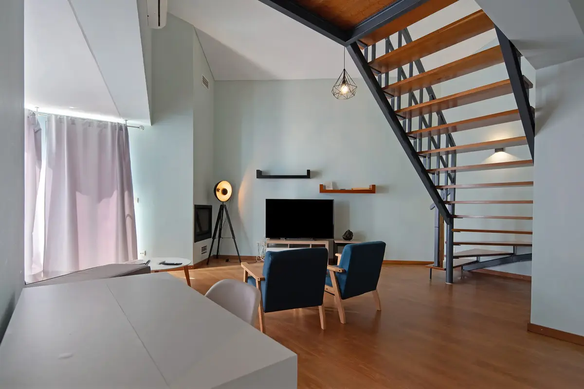  Spacious and modern open-plan living room at Ponta Delgada Apartment T3, featuring a dedicated white desk workstation, blue armchairs, a flat-screen TV, and an architectural wooden staircase
