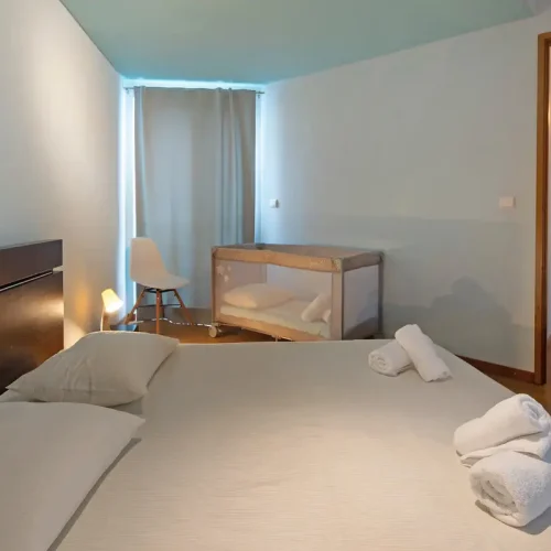 EN: Family bedroom at Ponta Delgada Apartment T3 featuring a double bed with white linens, a complimentary baby crib, a white chair, and a view into the hallway. PT: Quarto familiar no Ponta Delgada Apartment T3 com cama de casal, roupa de cama branca, berço de bebé gratuito, cadeira branca e vista para o corredor.