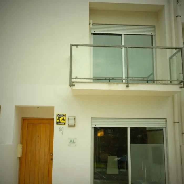 EN: Modern white facade of Guest House da Fajã de Baixo featuring a wooden entrance door, glass balcony, and large sliding windows in Ponta Delgada. PT: Fachada moderna e branca da Guest House da Fajã de Baixo com porta de entrada em madeira, varanda de vidro e grandes janelas de correr em Ponta Delgada.