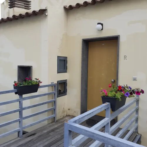 EN: Private entrance door of Ponta Delgada Apartment T3 on the first floor, featuring a wooden deck walkway, grey safety railings with flower boxes, and a stone door frame. PT: Porta de entrada privada do Ponta Delgada Apartment T3 no primeiro piso, com passadiço em deck de madeira, guardas de segurança cinzentas com floreiras e moldura de porta em pedra.