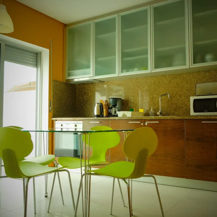 EN: Fully equipped modern kitchen with wooden cabinets, glass cupboards, microwave, and a glass dining table with lime green chairs at Guest House da Fajã de Baixo. PT: Cozinha moderna totalmente equipada com armários de madeira, vitrines de vidro, micro-ondas e mesa de jantar em vidro com cadeiras verde-lima na Guest House da Fajã de Baixo.