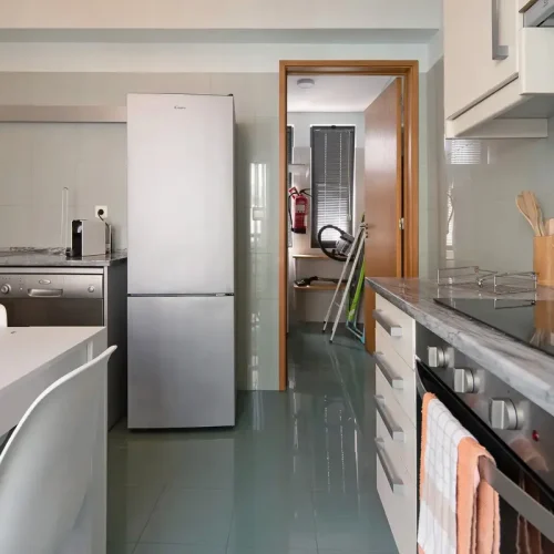 Fully equipped kitchen at Ponta Delgada Apartment T3 featuring stainless steel appliances, a grey tiled floor, and a view into a separate laundry room with a fire extinguisher. PT: Cozinha totalmente equipada no Ponta Delgada Apartment T3 com eletrodomésticos em aço inoxidável, chão de azulejo cinzento e vista para uma lavandaria separada com extintor.