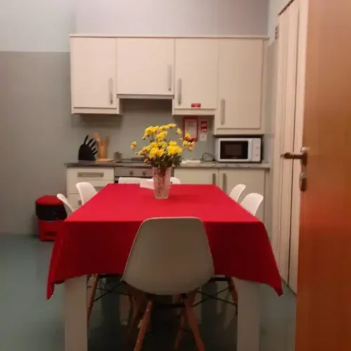 EN: Dining area and kitchenette at Ponta Delgada Apartment T3 featuring a table with a red tablecloth, white chairs, yellow flowers, and white kitchen cabinets with a microwave. PT: Área de refeições e kitchenette no Ponta Delgada Apartment T3 com mesa de toalha vermelha, cadeiras brancas, flores amarelas e armários de cozinha brancos com micro-ondas.