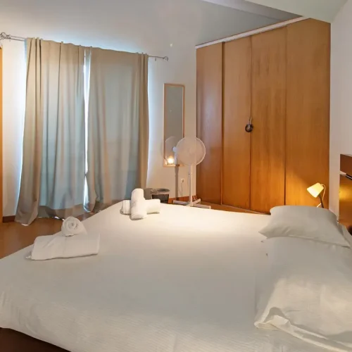 N: Spacious double bedroom at Ponta Delgada Apartment T3 featuring a large bed with white linens, wooden wardrobes, a floor fan, and warm bedside lighting. PT: Quarto de casal espaçoso no Ponta Delgada Apartment T3 com cama grande, roupa de cama branca, roupeiros em madeira, ventoinha de pé e iluminação de cabeceira quente.