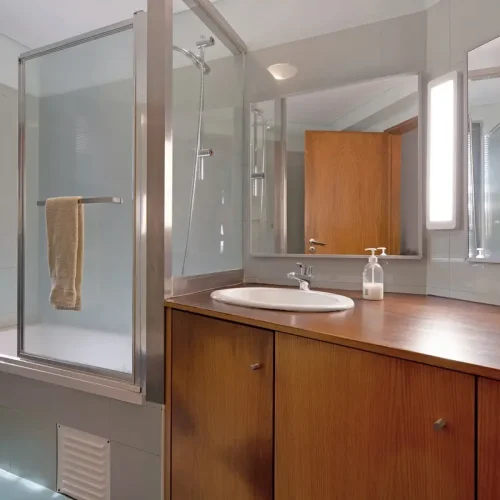 Modern bathroom at Ponta Delgada Apartment T3 featuring a bathtub with a glass shower screen, a large wooden vanity with a white sink, and two large mirrors. PT: Casa de banho moderna no Ponta Delgada Apartment T3 com banheira e resguardo de vidro, bancada larga em madeira com lavatório branco e dois espelhos grandes.