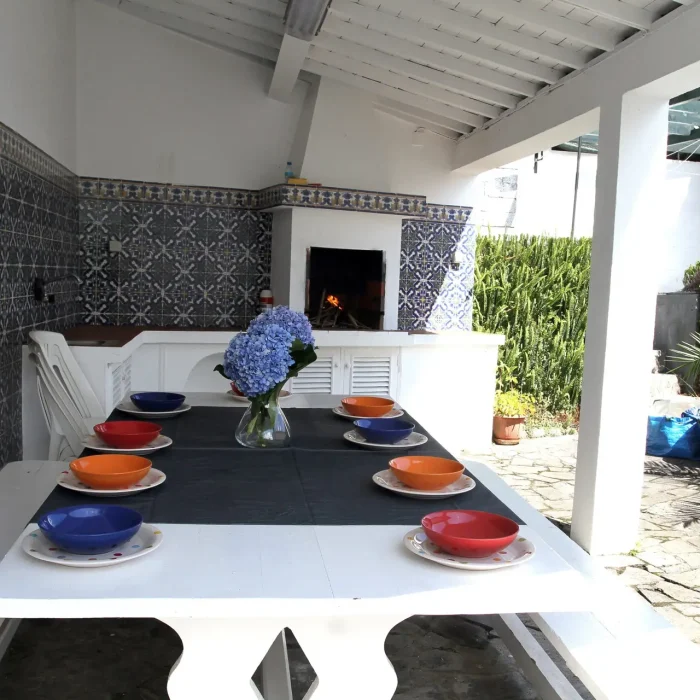Traditional outdoor dining area with barbecue, Portuguese blue tile walls, colorful dinnerware and garden access at São Miguel Azores vacation villa" Descrição: "Espaço exterior com churrasqueira tradicional, azulejos portugueses, perfeito para refeições em família em São Miguel