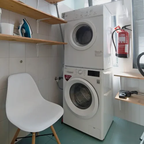 Fully equipped laundry room at Ponta Delgada Apartment T3 featuring a stacked washing machine and dryer, a steam iron, wooden shelving, and a fire extinguisher. PT: Lavandaria totalmente equipada no Ponta Delgada Apartment T3 com máquinas de lavar e secar roupa sobrepostas, ferro a vapor, prateleiras de madeira e extintor de incêndio.