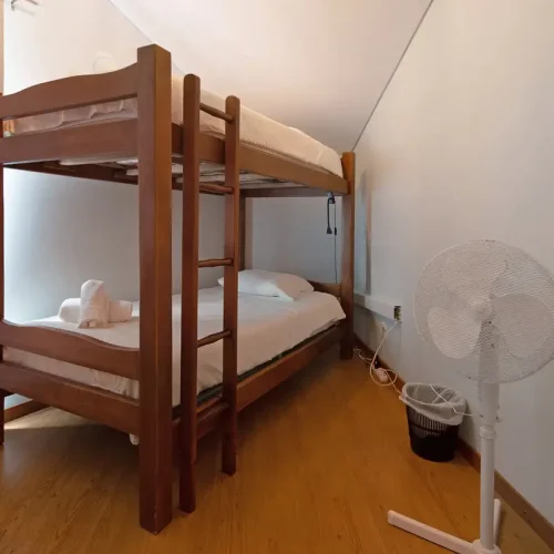 : Third bedroom of Ponta Delgada Apartment T3 on the upper floor, featuring wooden bunk beds with white linens, a white floor fan, and warm wooden flooring. PT: Terceiro quarto do Ponta Delgada Apartment T3 no piso superior, com beliches de madeira, roupa de cama branca, uma ventoinha de pé branca e chão de madeira acolhedor.