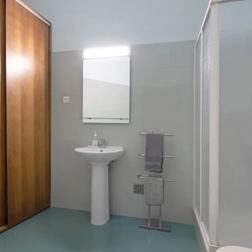 Full private bathroom on the upper floor of Ponta Delgada Apartment T3 featuring a glass shower cabin, white pedestal sink, large mirror with integrated lighting, and a wooden wardrobe. PT: Casa de banho privativa completa no piso superior do Ponta Delgada Apartment T3 com cabine de duche em vidro, lavatório de coluna branco, espelho grande com iluminação integrada e roupeiro em madeira.