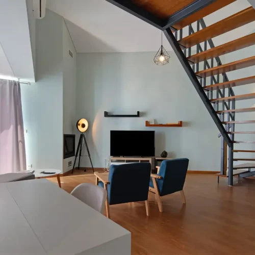 EN: Spacious and modern open-plan living room at Ponta Delgada Apartment T3, featuring a dedicated white desk workstation, blue armchairs, a flat-screen TV, and an architectural wooden staircase. PT: Sala de estar em plano aberto moderna e espaçosa no Ponta Delgada Apartment T3, com um posto de trabalho dedicado, poltronas azuis, TV de ecrã plano e uma escadaria de madeira arquitetónica.